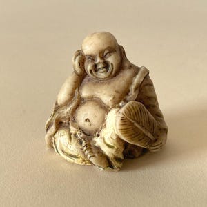 May include: A small, off-white, carved figurine of a laughing, seated figure with a bald head. The figure has one hand resting near the head and the other hand holding a textured, leaf-shaped fan. The surface features dark brown antiqued detailing.