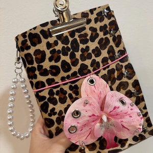 May include: A rectangular clipboard covered in a brown and black leopard print material, featuring a silver metal clip at the top, a pink elastic band, small silver star studs along the side, and a large pink orchid flower embellishment.