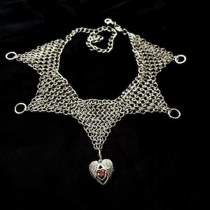 Könnte beinhalten: Eine silberfarbene Kettenhemd-Halskette mit sternförmigem Design und vier kreisförmigen Ringen an den Spitzen. Ein zentraler Anhänger in Form eines Herzens mit Flügeln und einem kleinen, facettierten roten Stein hängt an der Unterseite.