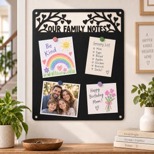 May include: A black rectangular magnetic board with the text "OUR FAMILY NOTES" and a decorative tree branch design with heart-shaped leaves at the top. The board holds four papers attached with colorful magnets, including a drawing, a list, and a photo.