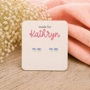 May include: A pair of small stud earrings shaped like sunglasses with horizontal stripes in yellow, blue, and pink. The earrings are attached to a square white display card with rounded corners that features the text made for Kathryn.