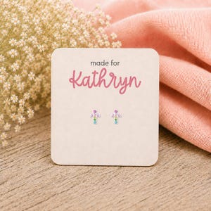 May include: A pair of small cross-shaped stud earrings featuring multicolored gemstones, displayed on a square beige card with the text "made for Kathryn" printed in pink. The earrings are silver-toned with pink, yellow, green, and blue stones.