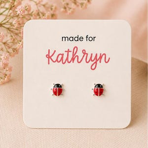 May include: A pair of silver stud earrings shaped like ladybirds with red and black enamel detailing. The earrings are attached to a square white display card featuring the black text made for and the pink script name Kathryn printed on the front.
