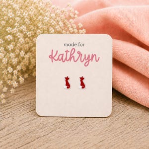 May include: A pair of small stud earrings shaped like foxes with red bodies and white tails. The earrings are attached to a square off-white display card featuring the text made for Kathryn in pink script lettering centered above the fox earrings.