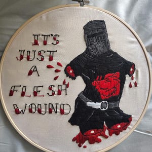 May include: A circular embroidery hoop containing a fabric piece with a black knight figure missing arms and legs, featuring red thread accents representing blood. The text IT'S JUST A FLESH WOUND is stitched in black with red blood-like details.