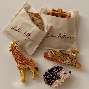 Peut inclure: Trois pinces à cheveux en forme d'animaux en résine translucide. Une pince représente une girafe jaune et orange, une autre un léopard tacheté et la troisième un hérisson marron et blanc. Deux sachets en papier beige portent l'inscription studio delfores.