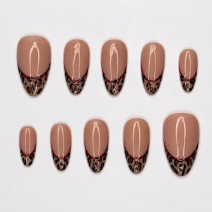 Puede incluir: Un juego de diez uñas postizas con una base de color nude y un diseño de manicura francesa. Las puntas presentan un estampado de leopardo marrón y negro, separado de la base nude por una fina línea horizontal de color rojo oscuro en cada uña.