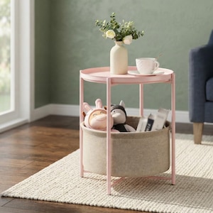 May include: A round light pink metal side table with a raised edge top and a lower beige fabric storage basket. The top surface holds a white ceramic vase with green foliage and a white textured teacup. Two plush toys sit inside the basket.