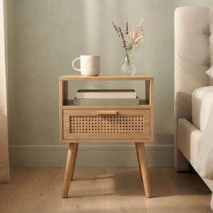 May include: A light wood nightstand featuring an open top shelf and a single drawer with a woven cane front panel. The nightstand stands on four tapered legs and holds a beige ceramic mug, a small glass vase with dried flowers, and two books.