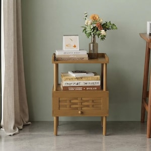 May include: A wooden nightstand with an open top shelf and a lower drawer featuring a woven front panel. The nightstand has four cylindrical legs and is topped with a glass vase of flowers, a small desk calendar, and stacked books.