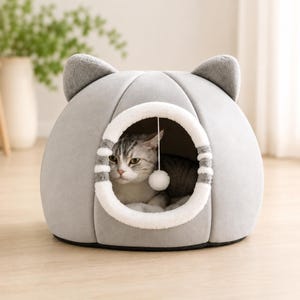Puede incluir: Una cama para mascotas en forma de cúpula de color gris claro con orejas de gato de felpa en la parte superior. La entrada circular presenta un borde blanco y difuso con detalles grises y un juguete de pompón blanco colgante.