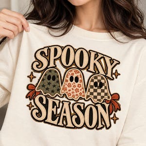 Może przedstawiać: Kremowy t-shirt z nadrukiem graficznym z napisem SPOOKY SEASON pogrubioną, ciemną czcionką. Wzór przedstawia trzy duszki z różnymi motywami: jeden w gwiazdki, drugi w kwiaty, a trzeci w szachownicę.