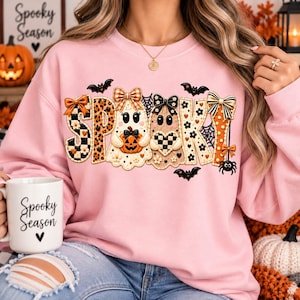Puede incluir: Una sudadera rosa claro con el texto SPOOKY en una fuente decorativa con patrones, lazos, fantasmas, calabazas, murciélagos y arañas. La sudadera se acompaña de una taza blanca con el texto Spooky Season y un corazón impreso.
