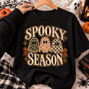 Könnte beinhalten: Ein schwarzes Kurzarm-T-Shirt mit dem Text SPOOKY SEASON in cremefarbener Schrift. Unter dem Text befinden sich drei Geisterillustrationen mit verschiedenen Mustern: einer mit grünen Sternen, einer mit orangefarbenen Blumen und einer mit schwarz-weißem Schachbrettmuster.