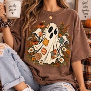May include: A brown short-sleeved t-shirt featuring a central graphic of a white ghost adorned with various colourful patterned patches, surrounded by orange and white autumn flowers. The ghost has large black eyes and a wide open mouth.