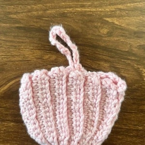 May include: A light pink crocheted ornament shaped like a seashell with vertical ribbed texture and a small looped yarn handle at the top for hanging. The item is crafted from soft yarn and features a scalloped edge along the bottom border.