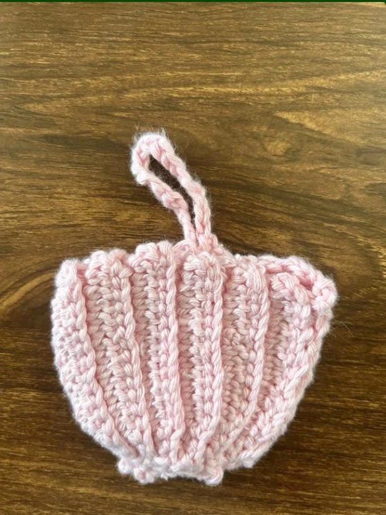 May include: A light pink crocheted ornament shaped like a seashell with vertical ribbed texture and a small looped yarn handle at the top for hanging. The item is crafted from soft yarn and features a scalloped edge along the bottom border.