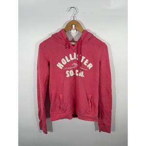 May include: A bright pink long-sleeve hooded sweatshirt featuring a white embroidered Hollister So. Cal logo with a wave graphic on the chest. The hoodie includes a drawstring neckline, ribbed cuffs, and two front pockets on a wooden hanger.