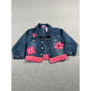 May include: A blue denim jacket featuring a pink fabric hem, a large pink flower appliqué on the right side, and a smaller pink flower patch on the left. The jacket includes silver-toned metal buttons, a classic collar, and long sleeves.