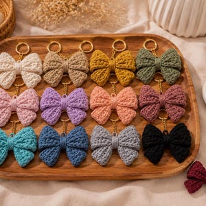 Könnte beinhalten: Eine Sammlung von dreizehn gehäkelten Schleifen-Schlüsselanhängern in verschiedenen Farben, darunter Creme, Beige, Senfgelb, Olivgrün, Hellrosa, Lavendel, Pfirsich, Mauve, Türkis, Blau, Grau, Schwarz und Dunkelrot, jeweils an einem goldfarbenen Metallring.