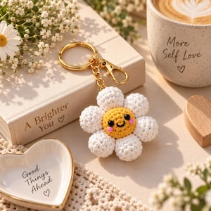 May include: A crochet daisy keychain featuring a yellow center with a smiling face and six white petals. The keychain includes a gold-toned metal ring and a lobster clasp. The center has black bead eyes, a black stitched mouth, and pink cheeks.