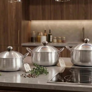 May include: A set of three brushed aluminium pots with matching lids and side handles. Each pot features a small white rectangular tag with the letter G attached to the handle. The pots are arranged in a row on a smooth countertop surface.