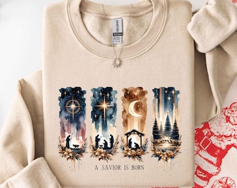 May include: A beige crewneck sweatshirt featuring a graphic print of four vertical panels depicting a nativity scene, stars, a crescent moon, and pine trees, with the text A SAVIOR IS BORN printed in black lettering below the illustrations.