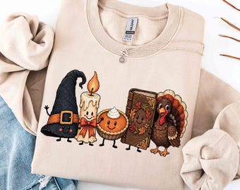 May include: A beige crewneck sweatshirt featuring a graphic print of a black witch hat, a lit candle, a pumpkin pie, a book with a wreath, and a turkey. The interior neck label displays the text Gildan.