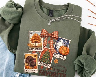 May include: An olive green crewneck sweatshirt featuring a graphic of four postage stamps representing September, October, November, and December. The stamps show a tree, a ghost with a pumpkin, a turkey, and a Christmas tree, tied with a plaid bow.