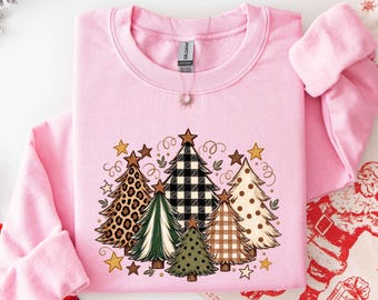 May include: A light pink crewneck sweatshirt featuring a graphic print of five stylized Christmas trees. The trees display various patterns including leopard print, black and white buffalo plaid, solid green, brown plaid, and polka dots.