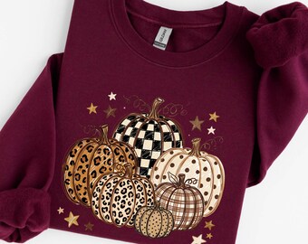 May include: A maroon crewneck sweatshirt featuring a graphic print of five pumpkins with various patterns including leopard print, black and white checkered, and polka dots, accented by small gold and white stars scattered around the design.