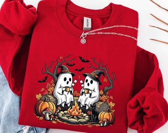 May include: A red crewneck sweatshirt featuring a graphic of two white ghosts wearing black witch hats, sitting on a log by a campfire. The design includes pumpkins, mushrooms, a black cat, autumn leaves, bare trees, and flying bats.