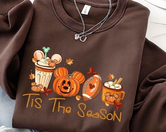 May include: A dark brown crewneck sweatshirt featuring a graphic print of a coffee cup, a pumpkin with a mouse-ear silhouette, a football, and a mug with marshmallows. The text "Tis The Season" is printed in orange below the graphic elements.