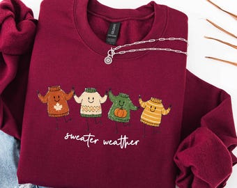 May include: A maroon crewneck sweatshirt featuring a graphic of four anthropomorphic sweaters holding hands. The sweaters are orange with a maple leaf, cream with a chevron pattern, green with a pumpkin, and yellow striped. The text reads sweater weather.