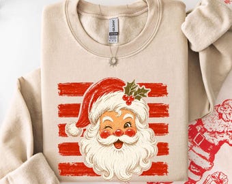 May include: A sand-colored crewneck sweatshirt featuring a graphic print of a winking Santa Claus face with a white beard, red hat, and holly berries. The design is set against a background of horizontal red brushstrokes on the front chest.