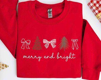 May include: A bright red long-sleeve sweatshirt featuring a white graphic print of three bows and two Christmas trees arranged in a row. Below the graphic, the text "merry and bright" is printed in a white, cursive, handwritten-style font.