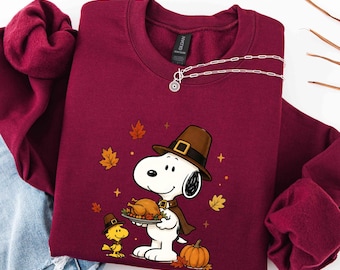May include: A maroon crewneck sweatshirt featuring a graphic of Snoopy and Woodstock wearing pilgrim hats. Snoopy holds a platter with a roasted turkey, surrounded by orange autumn leaves and small stars. The Gildan brand tag is visible.