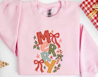 May include: A light pink crewneck sweatshirt featuring a colorful graphic print of the word Merry. The letters are decorated with red, pink, green, and yellow patterns, accompanied by illustrations of red bows, holly leaves, and a candy cane.