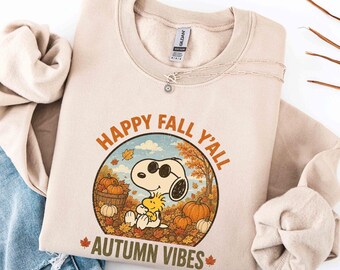 May include: A beige crewneck sweatshirt featuring a graphic of Snoopy wearing sunglasses and holding Woodstock amidst pumpkins and autumn leaves. The text reads HAPPY FALL Y'ALL and AUTUMN VIBES in orange and brown lettering.