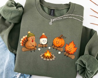 May include: An olive green crewneck sweatshirt featuring a graphic of a turkey, a marshmallow, a pumpkin, and an autumn leaf sitting on logs around a campfire. Each character holds a stick with a marshmallow over the orange and yellow flames.