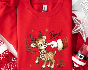 May include: A red crewneck sweatshirt featuring a graphic of a brown reindeer wearing a red and white striped scarf. A hand in a red and white Santa sleeve is touching the reindeer's nose, with the text Boop written in black script above.