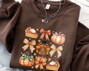 May include: A dark brown crewneck sweatshirt featuring a graphic print of autumn-themed icons including pumpkin pie slices, various patterned bows, pumpkins, and a turkey wearing a pilgrim hat, accented by a silver chain necklace.
