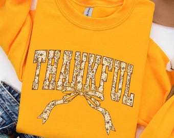 May include: A bright yellow crewneck sweatshirt featuring the word THANKFUL in large block letters. The letters and a decorative bow graphic below the text are filled with a cream-colored pattern of small brown flowers and green leaves.