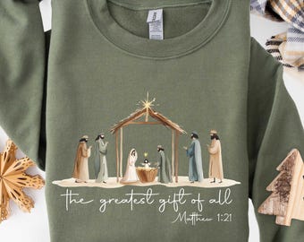 May include: An olive green crewneck sweatshirt featuring a printed nativity scene illustration with a stable, figures, and a star. Below the scene, the text reads: the greatest gift of all Matthew 1:21 in a white cursive and sans-serif font.