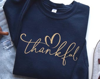 May include: A navy blue crewneck sweatshirt featuring the word thankful written in a gold cursive script font with a heart shape integrated into the top of the letter h. The garment has long sleeves and a ribbed collar, showcasing a clean design.