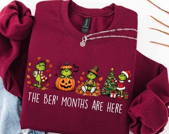 May include: A maroon crewneck sweatshirt featuring a graphic of a green character in four seasonal scenes, with the text "THE BER' MONTHS ARE HERE" printed in white below the illustrations. The sweatshirt has a Gildan brand label inside.