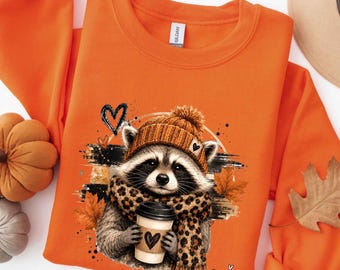 May include: An orange crewneck sweatshirt featuring a graphic of a raccoon wearing a brown knit beanie and a leopard print scarf. The raccoon holds a coffee cup with a heart design. The print includes autumn leaves and black heart accents.