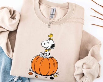 May include: A beige crewneck sweatshirt featuring a graphic of Snoopy sitting on a large orange pumpkin with Woodstock perched on Snoopy's head. The design includes small autumn leaves and an acorn at the base. The garment has a Gildan brand label.