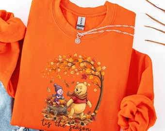 May include: An orange crewneck sweatshirt featuring a graphic of Winnie the Pooh and Piglet with a pumpkin in a wooden cart under an autumn tree. The text "tis the season" is printed below the illustration in a black cursive script font.