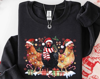 May include: A black long-sleeve sweatshirt featuring a graphic of three chickens wearing red and white Santa hats. The center chicken wears a red and white striped scarf. The chickens are decorated with colorful Christmas lights, surrounded by gift boxes, candy canes, and snowflakes.